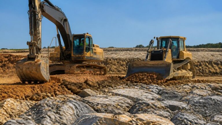 Escavadeira e bulldozer operando em terreno variado com áreas de solo arenoso e rochoso